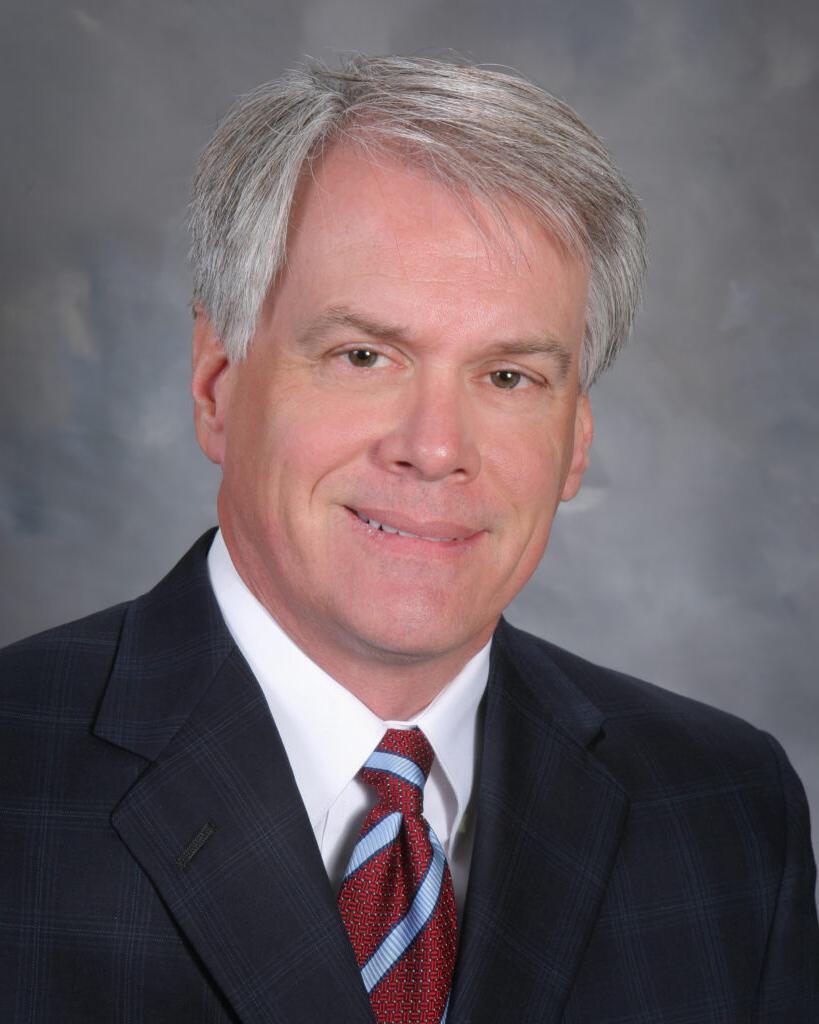 A man with gray hair wearing a dark suit, white shirt, and red-striped tie, poses against a neutral backdrop.